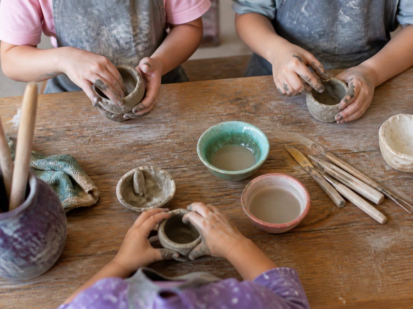 Three children in aprons shaping clay pinch pots at a wooden table, tools and water bowls between them