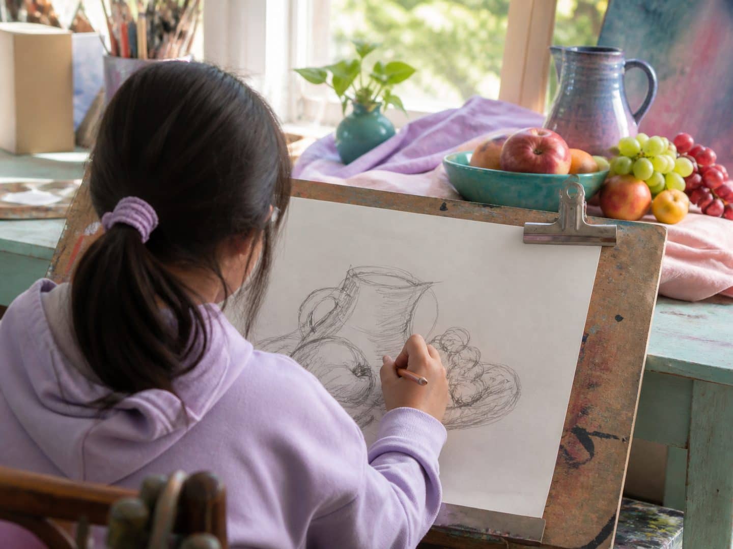 A child drawing a still life in pencil, with a jug and a bowl of fruit set up on the table