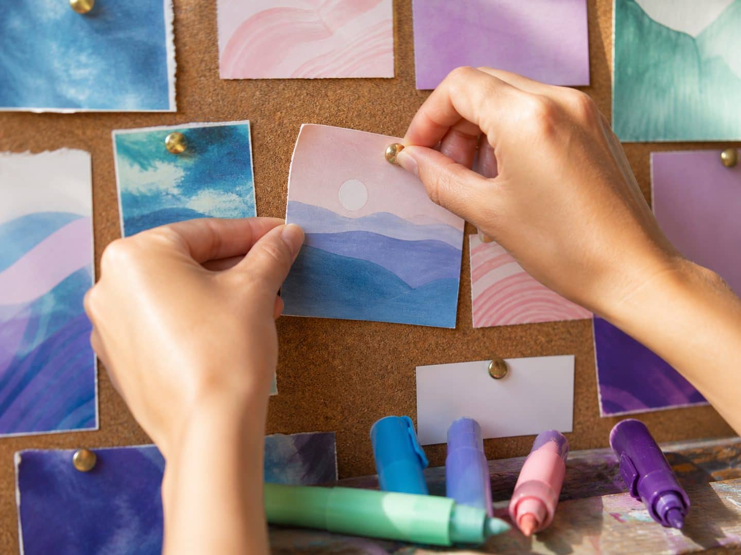 Two hands pinning a painted card onto a cork board already covered in watercolour swatches